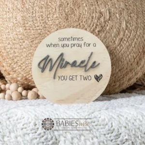 Round timber twin pregnancy announcement plaque featuring the phrase :sometimes when you pray for a Miracle, you get two" with the word "Miracle" in raised 3D acrylic . Personlised with the surname, month and year UV printed along the bottom of the plaque. Styled on a soft white blanket with a woven pillow & wooden beads.