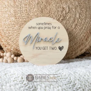 Round timber twin pregnancy announcement plaque featuring the phrase :sometimes when you pray for a Miracle, you get two" with the word "Miracle" in raised 3D acrylic . Personlised with the surname, month and year UV printed along the bottom of the plaque. Styled on a soft white blanket with a woven pillow & wooden beads.