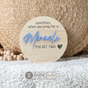 Round timber twin pregnancy announcement plaque featuring the phrase :sometimes when you pray for a Miracle, you get two" with the word "Miracle" in raised 3D acrylic . Personlised with the surname, month and year UV printed along the bottom of the plaque. Styled on a soft white blanket with a woven pillow & wooden beads.