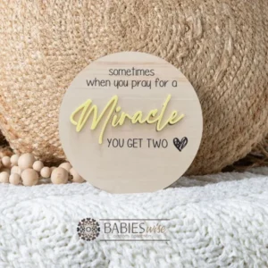 Round timber twin pregnancy announcement plaque featuring the phrase :sometimes when you pray for a Miracle, you get two" with the word "Miracle" in raised 3D acrylic . Personlised with the surname, month and year UV printed along the bottom of the plaque. Styled on a soft white blanket with a woven pillow & wooden beads.