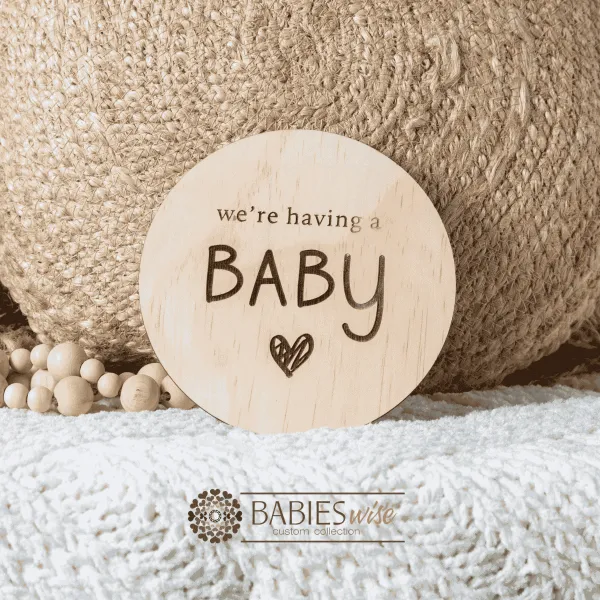 Round timber pregnancy announcement plaque with the words "we're having a baby" engraved in dark letters. Styled on a soft white blanket with a woven pillow & wooden beads.